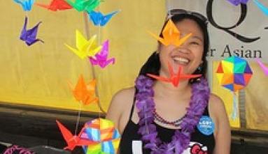 Woman in Hello Kitty "LOVE" tank top stands behind a table draped with rainbow origami cranes at Boston Pride